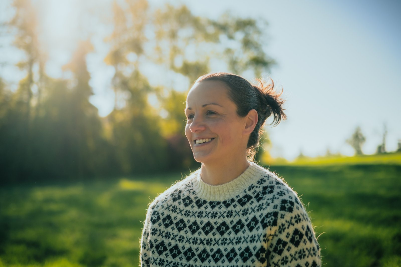 Nadine Vergote steht lächelnd im Strickpullover auf einer sonnigen Wiese vor Bäumen